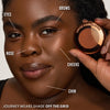 Eine Frau hält eine Dose Yummy Skin Blurring Balm Powder Lowlighter Off The Grid neben ihr Gesicht lächelt. 