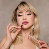 Eine Frau mit blonden Haaren posiert mit dem Lip Frame Pencil vor einer grau-beigen Wand. Die Farbe Cool Girl Pink auf den Lippen.