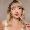 Eine Frau mit blonden Haaren posiert mit dem Lip Frame Pencil vor einer grau-beigen Wand. Die Farbe Soft Brownie auf den Lippen.