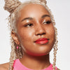 Lächelnde Frau mit mittlerem Hautton, rotem Lippenstift und blondierten Braids präsentiert ihren strahlenden Look mit MAGIC FILTER Glow Booster 40 Tan.