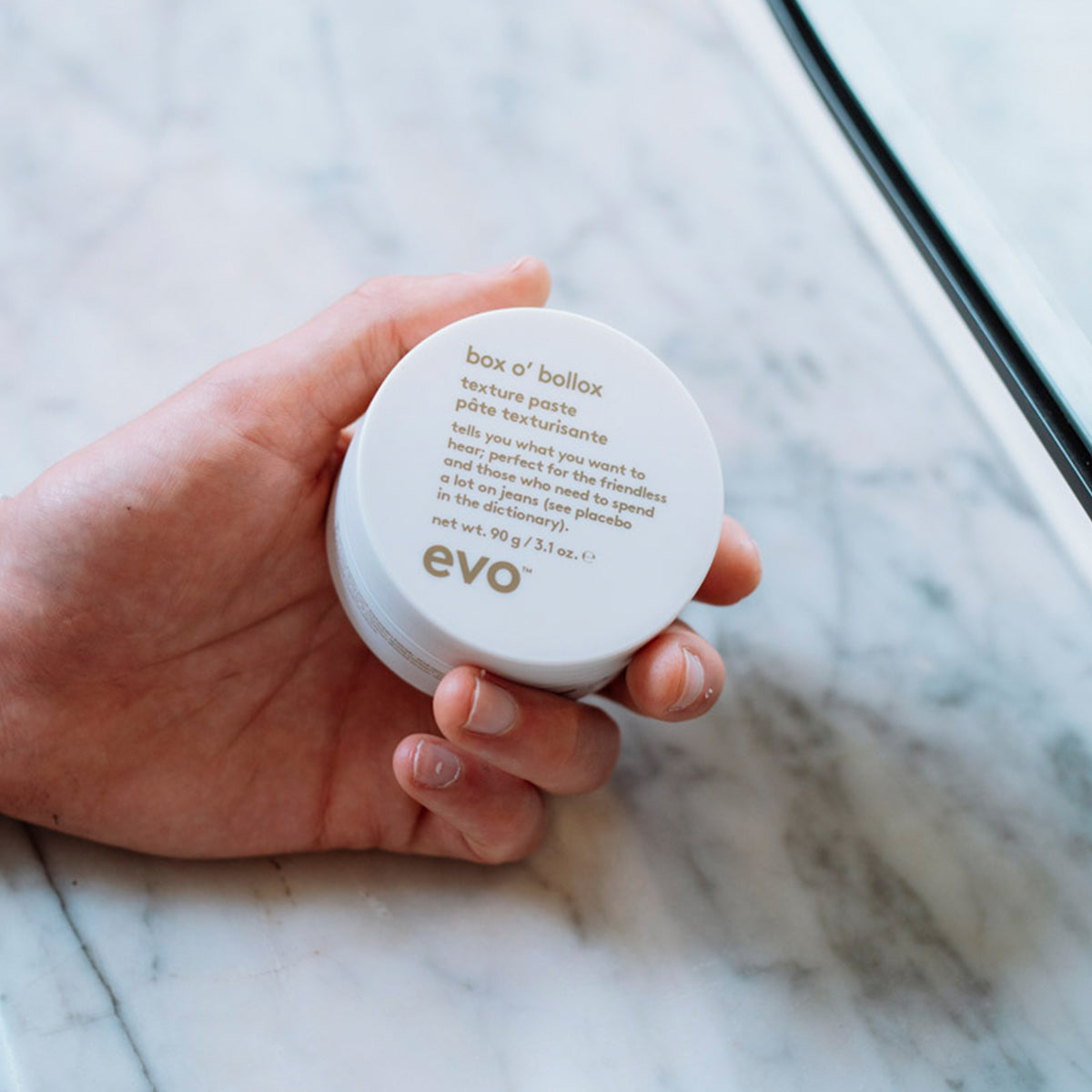 Eine Hand hält einen runden weißen Behälter Box O Bollox Texture Paste auf einer Marmoroberfläche.
