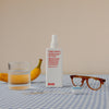 Eine Flasche Happy Campers Wearable Treatment, ein Glas Wasser, eine Banane, eine Brille und eine Zahnbürste auf einer blau karierten Tischdecke.