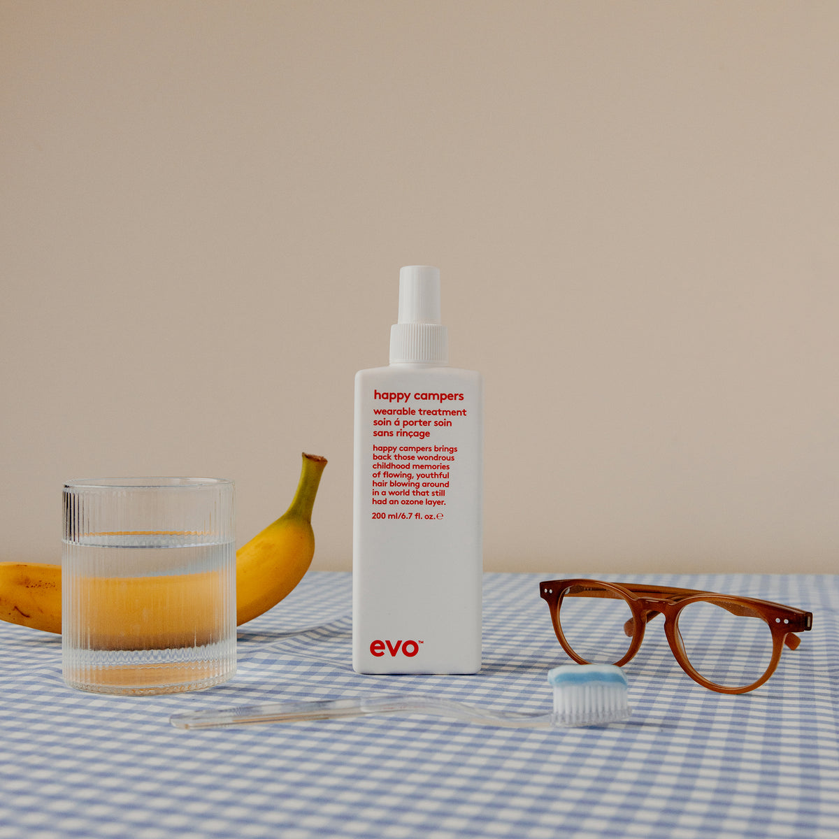 Eine Flasche Happy Campers Wearable Treatment, ein Glas Wasser, eine Banane, eine Brille und eine Zahnbürste auf einer blau karierten Tischdecke.