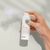 Eine Hand sprüht Haze Styling Powder aus einer weißen Flasche vor einem hellen Hintergrund.