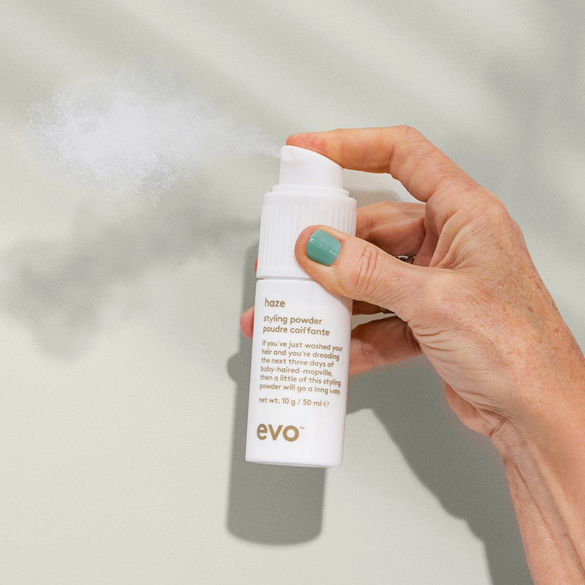 Eine Hand sprüht Haze Styling Powder aus einer weißen Flasche vor einem hellen Hintergrund.