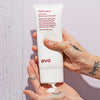 Eine Hand verteilt Liquid Rollers Curl Balm auf der Handfläche vor einer violetten Wand.