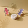 Zwei gestreifte Miniatur-Liegestühle und ein Tiegel The Shag Beach Paste auf einer gelben Oberfläche mit Schatten von Palmen.