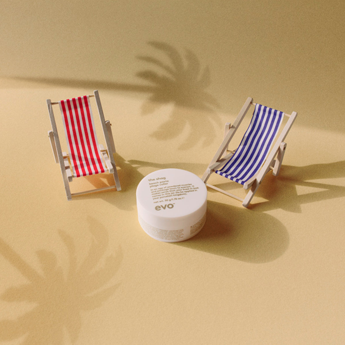 Zwei gestreifte Miniatur-Liegestühle und ein Tiegel The Shag Beach Paste auf einer gelben Oberfläche mit Schatten von Palmen.