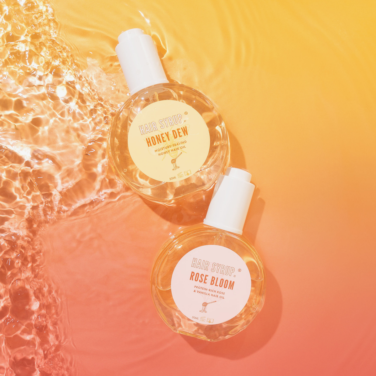 Zwei Hair-Syrup-Haaröle, Honey Dew und Rose Bloom, schwimmen im Wasser auf gelb-rosa Farbverlauf, Etiketten sichtbar.