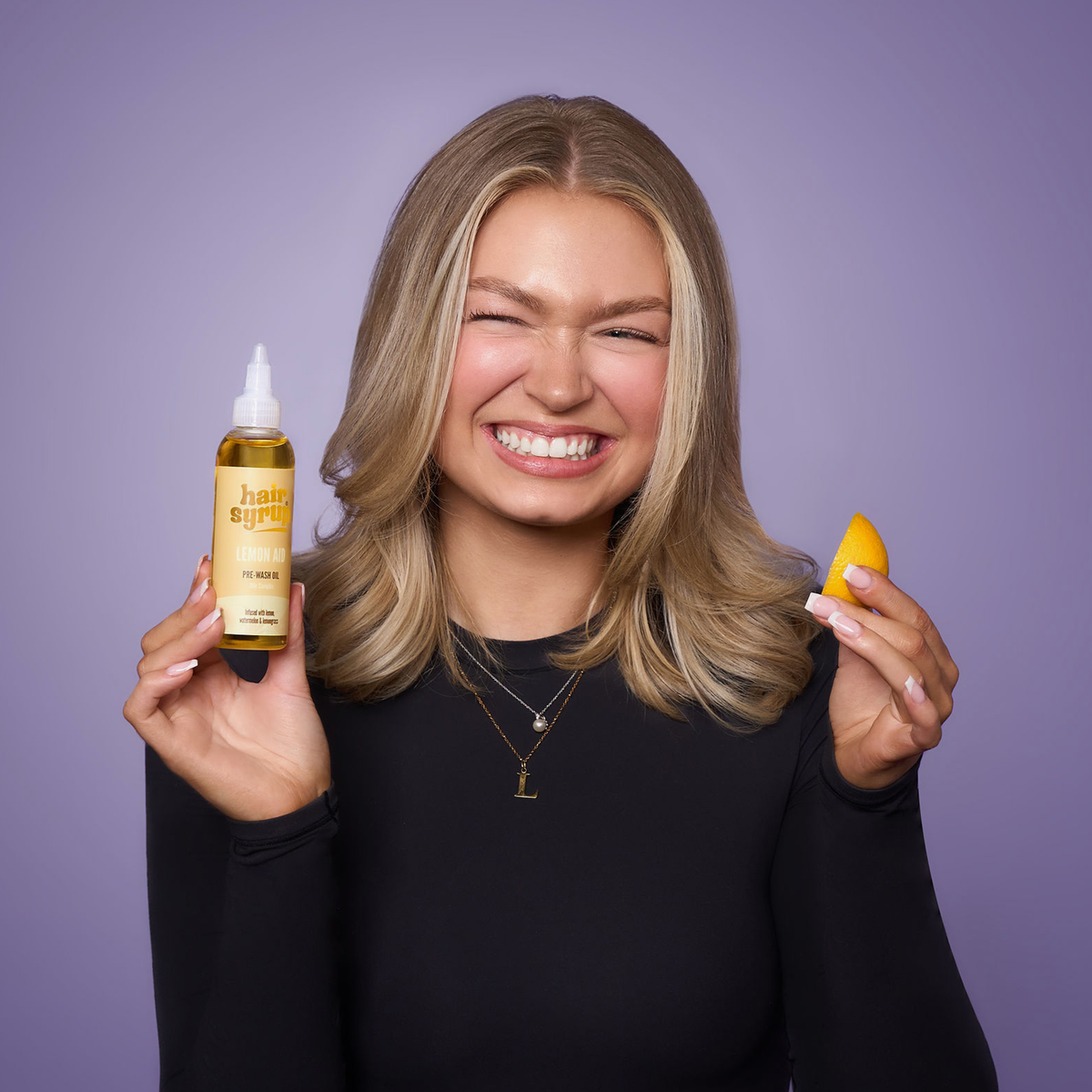 Eine lächelnde Frau hält eine Flasche Vitamin C Me-Haaröl von Hair Syrup in der einen und eine Orangenspalte in der anderen Hand vor einem lila Hintergrund.