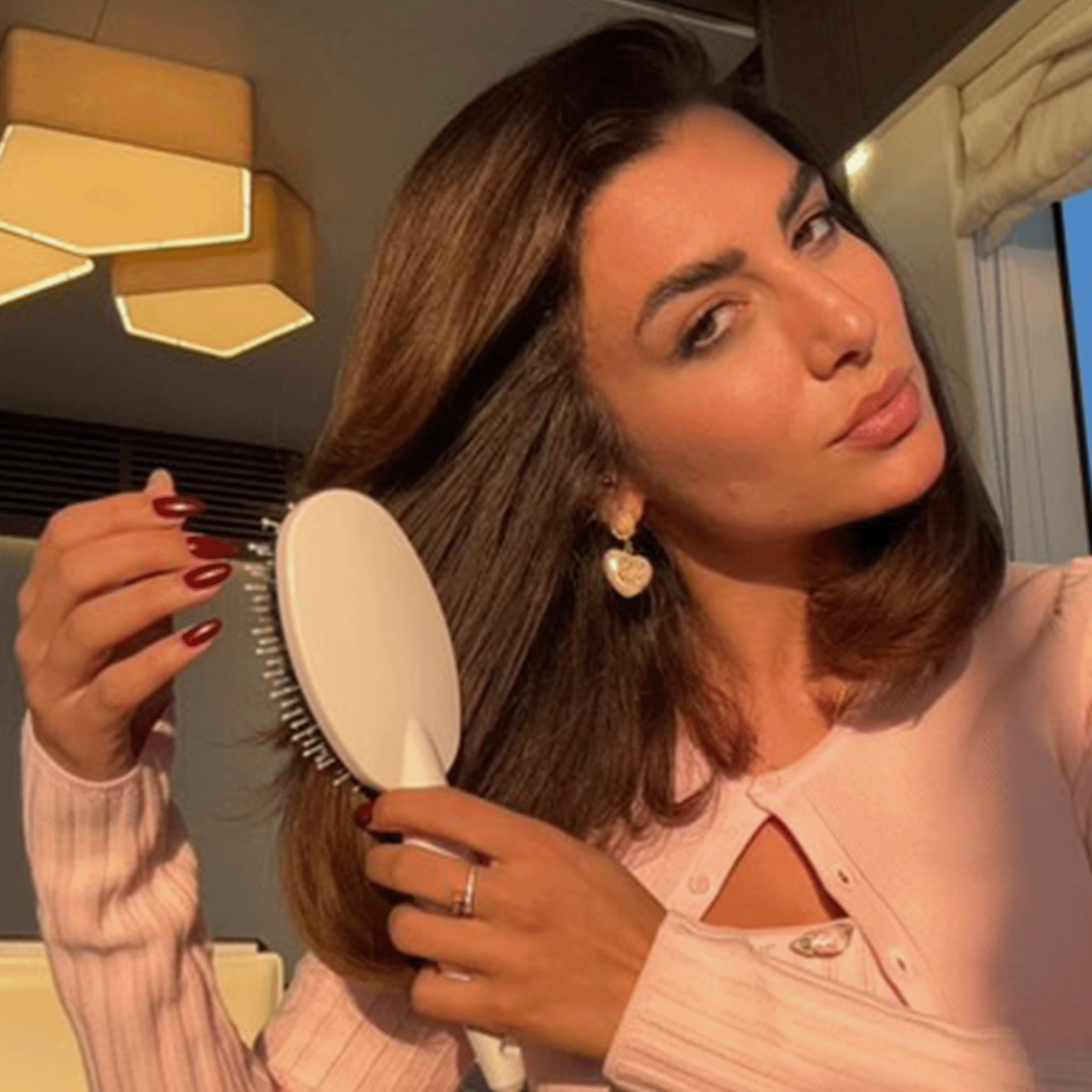 Frau mit braunen Haaren bürstet ihr Haar mit der weißen Miracle Brush x Hairtamin in warmem Innenraumlicht.