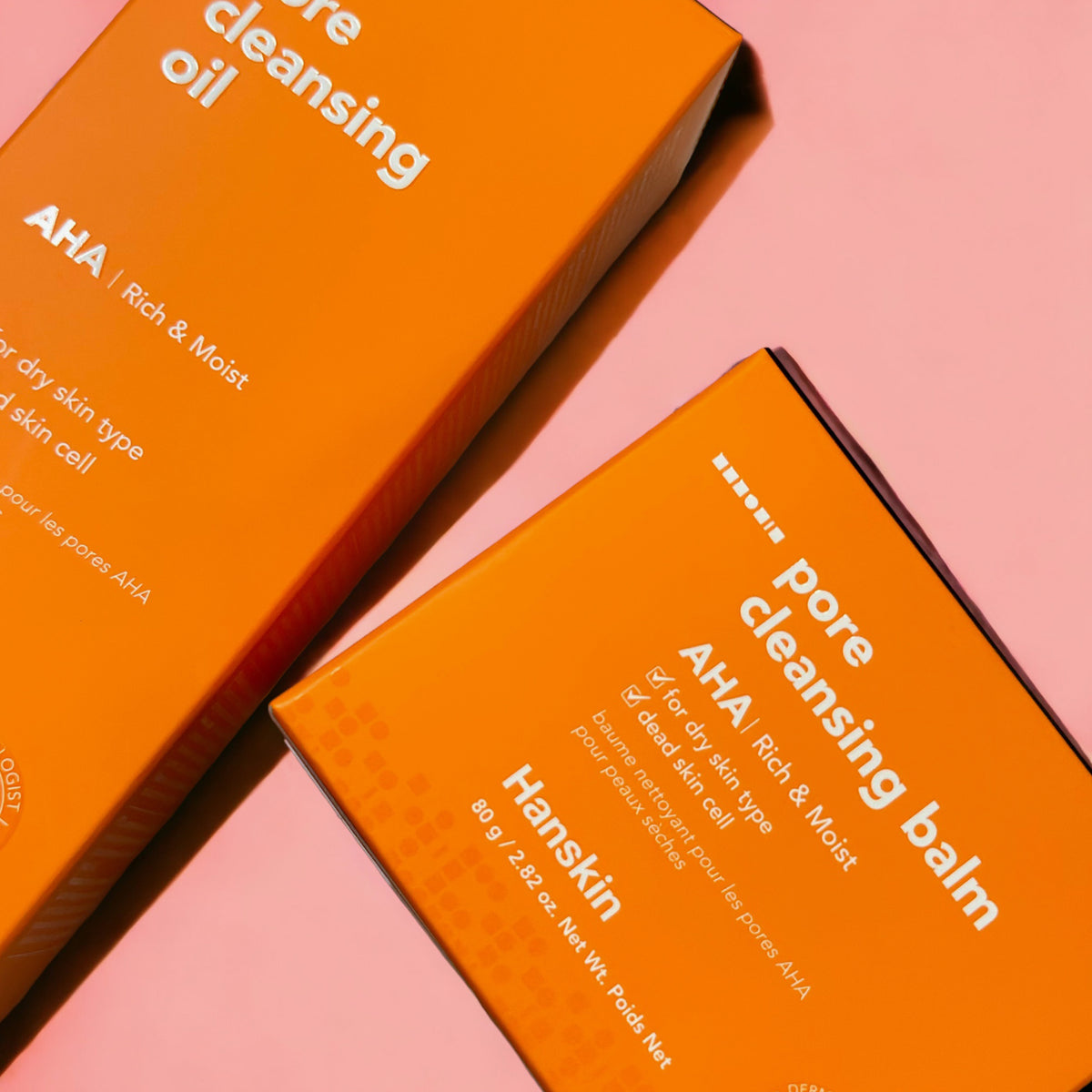 Zwei orangefarbene Hanskin-Boxen mit der Aufschrift „Pore Cleansing Oil AHA“ und „Pore Cleansing Balm AHA“ auf rosa Hintergrund.