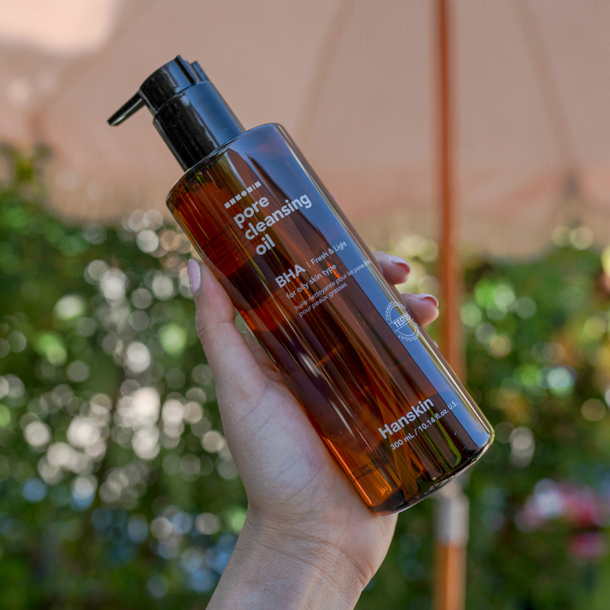 Eine Hand hält im Freien eine Flasche Pore Cleansing Oil BHA, im Hintergrund ist Grün.