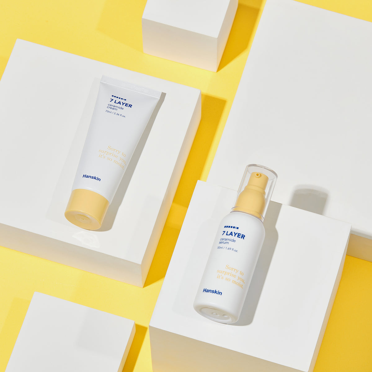 Das Hanskin Seven Layer Ceramide Serum und die Cream auf weißen Blöcken vor gelbem Hintergrund.