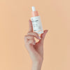 Eine Hand hält eine Flasche Vitamin C Glow Serum vor einem pfirsichfarbenen Hintergrund.