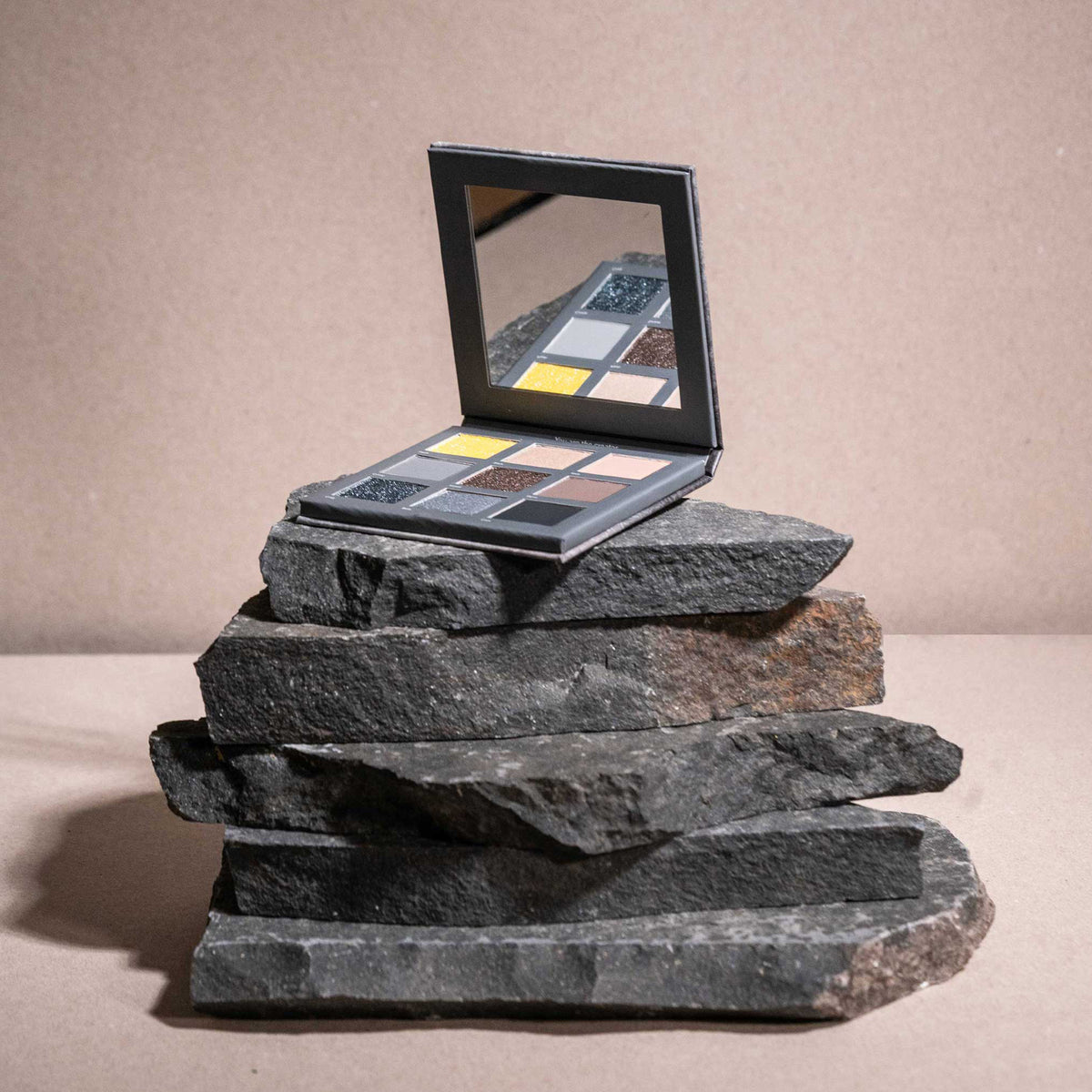 Die Aim Higher Palette von LH Cosmetics mit einer Reihe von Lidschattentönen liegt offen auf gestapelten dunklen Steinen mit beigem Hintergrund.