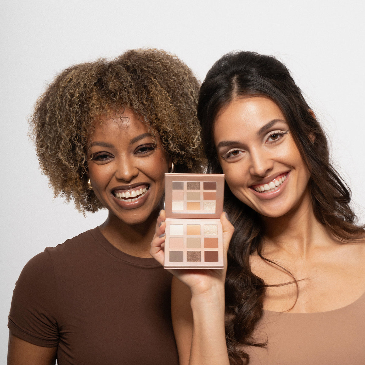 Zwei lächelnde Frauen, eine hält die Boring Nude Palette von LH Cosmetics – eine offene Lidschattenpalette mit neutralen Farbtönen – vor einem weißen Hintergrund.