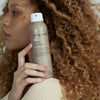 Frau mit lockigem blondiertem Haar hält eine Flasche Living Proof No Frizz Instant De-Frizzer vor ihr Gesicht.