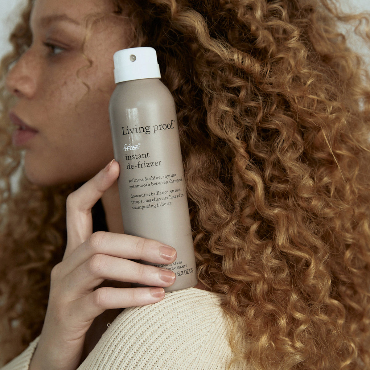 Frau mit lockigem blondiertem Haar hält eine Flasche Living Proof No Frizz Instant De-Frizzer vor ihr Gesicht.