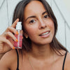 Eine Frau mit langen braunen Haaren lächelt in die Kamera und hält eine Flasche Lumene NORDIC-C [VALO] Arctic Berry Multivitamin Cocktail Oil.