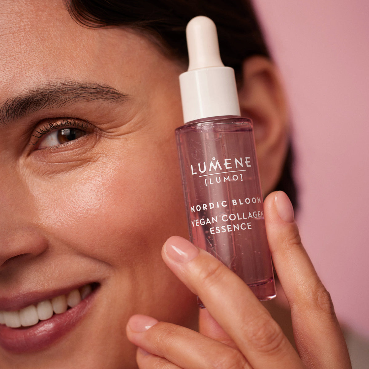 Eine lächelnde Person hält eine Flasche Lumene NORDIC BLOOM [LUMO] Vegan Collagen Essence an ihre Wange.