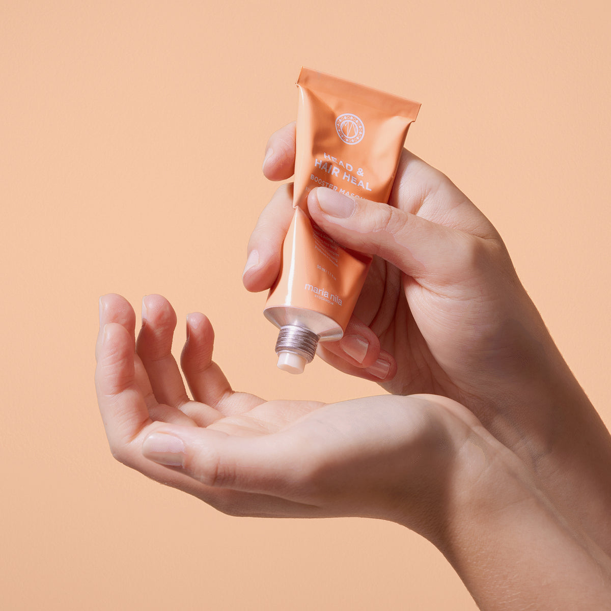 Vor einem pfirsichfarbenen Hintergrund drückt eine Person die maria nila Booster Masque Head & Heal aus einer orangefarbenen Tube auf ihre Hand.