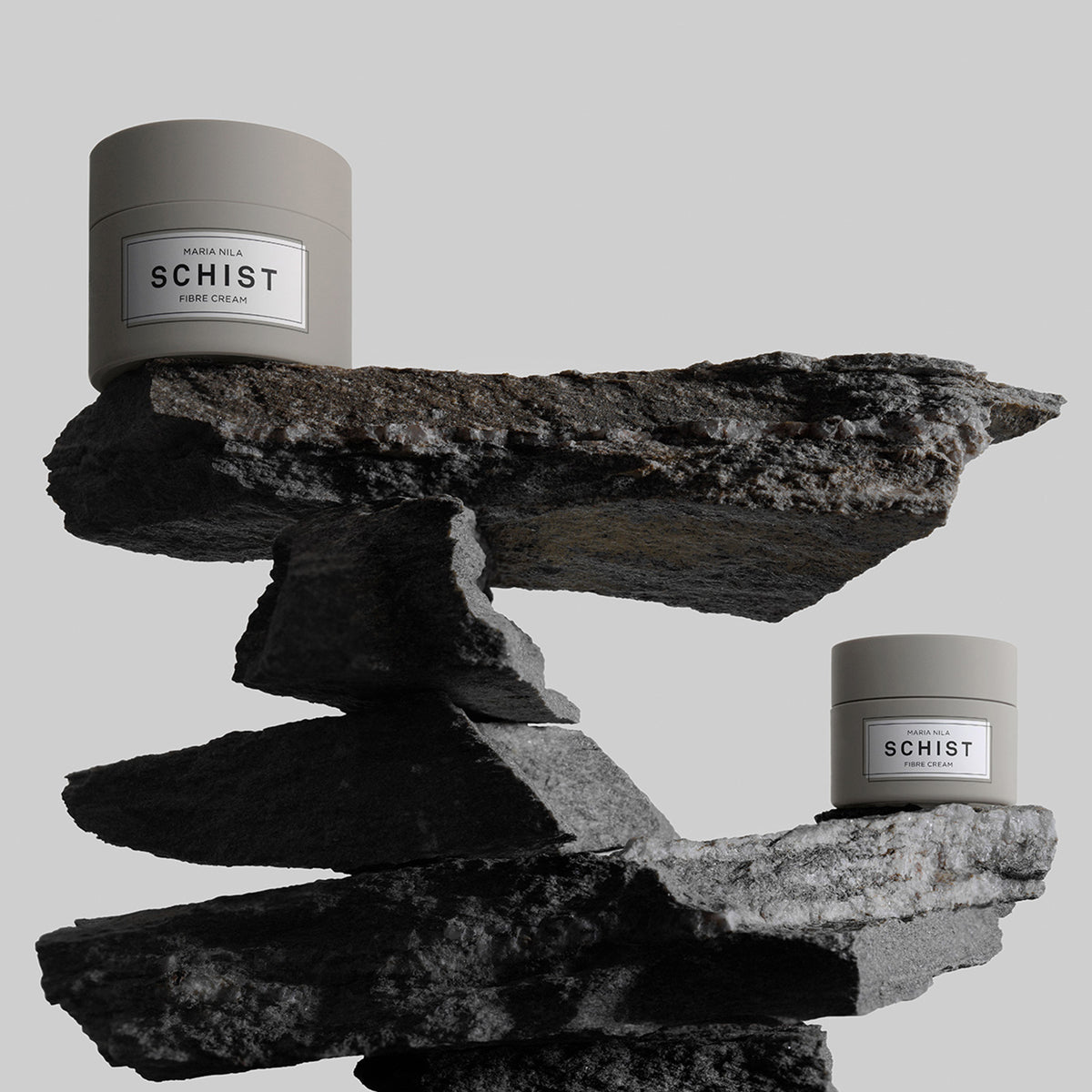 Zwei graue Tiegel der Schist Fibre Cream von maria nila stehen auf gestapelten, ungleichmäßigen dunklen Steinen.