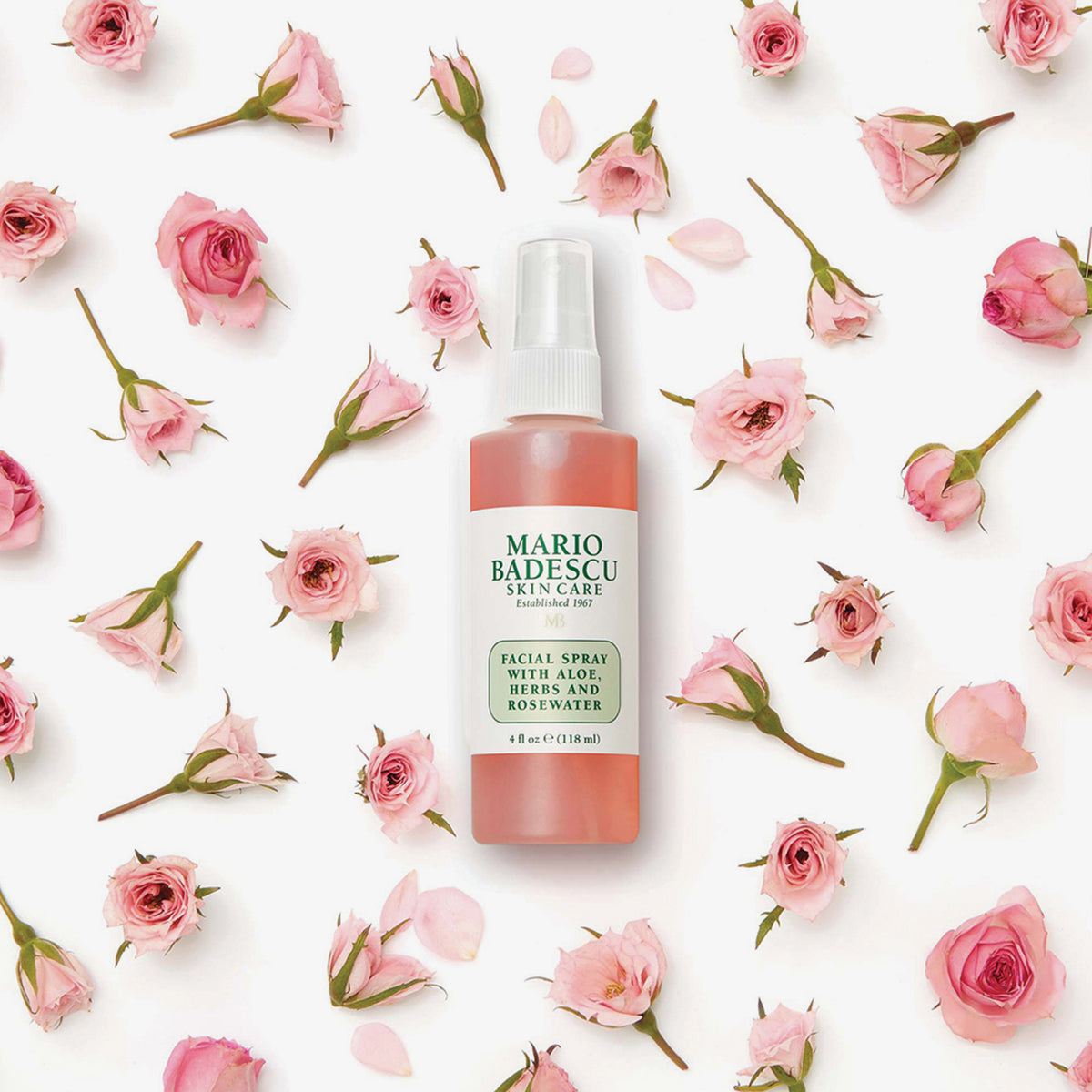 Eine Flasche Mario Badescu Gesichtsspray mit Aloe, Kräutern und Rosenwasser ist von verstreuten rosa Rosen auf weißem Hintergrund umgeben.