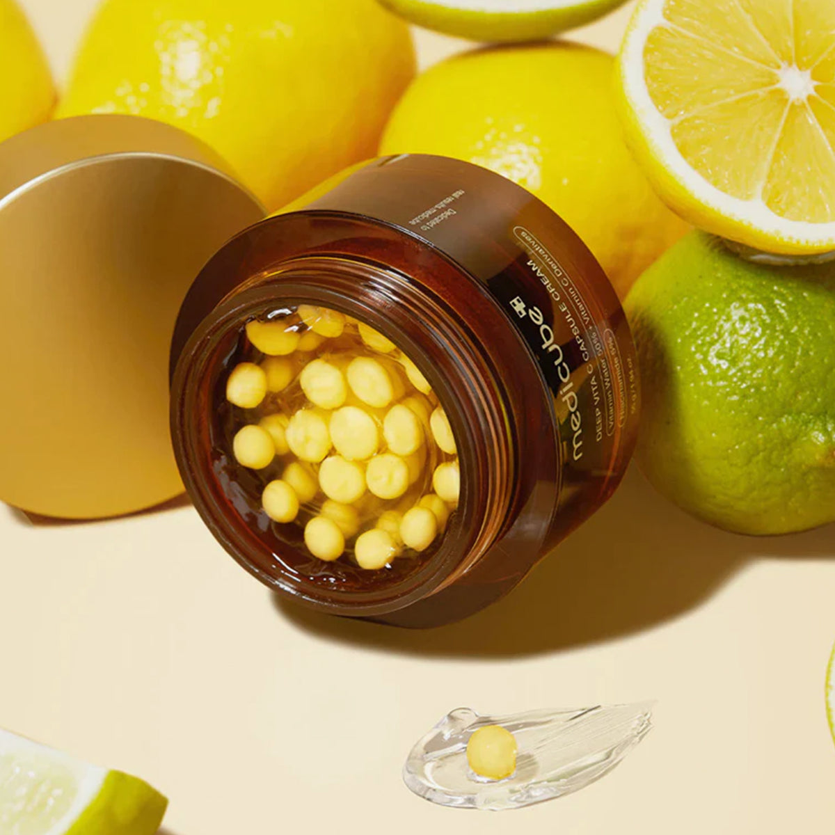 Offenes Glas Deep Vita C Capsule Cream mit gelben Kapseln, umgeben von frischen Zitronen und Limetten auf einer hellen Oberfläche.