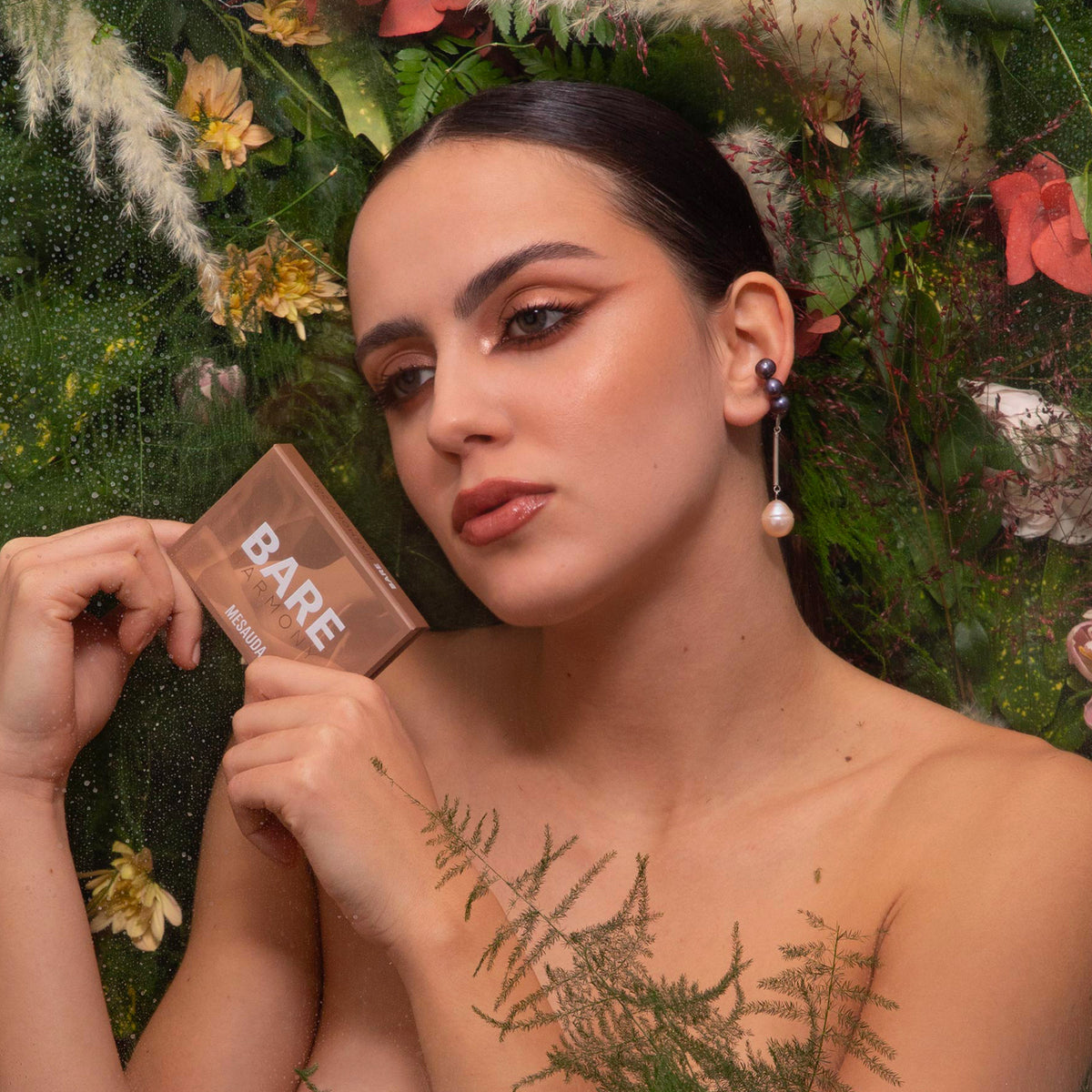 Eine Frau mit kräftigem Make-up hält die Mesauda Beauty Bare Harmony 3.0 Palette in der Hand, umgeben von Blumen und Grünzeug.