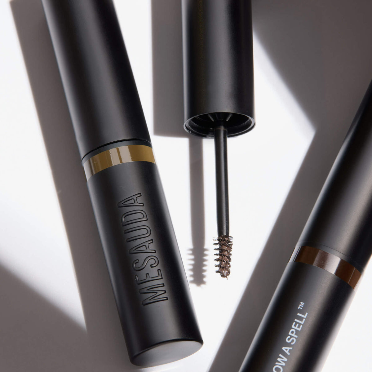 Drei Tuben Brow a Spell Augenbrauengel von Mesauda Beauty, von denen eine geöffnet ist, um einen mit braunem Gel beschichteten Pinsel zu enthüllen.