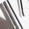 Eine silberne Mesauda Beauty Brow Glaminate - 101 Clear Tube mit einem gebogenen Borstenkopf daneben auf weißem Hintergrund.