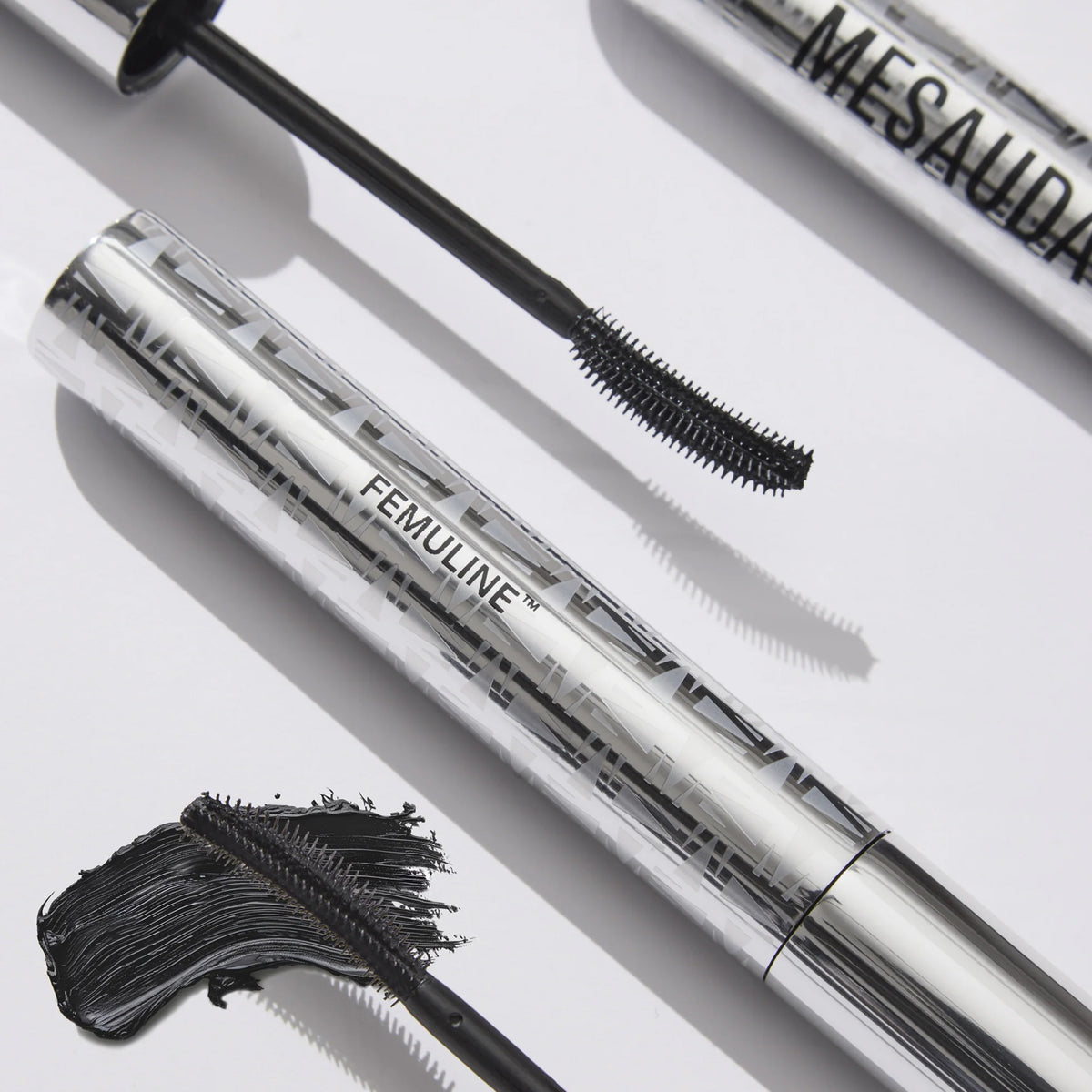 Zwei silberne Wimperntusche-Tuben mit der Aufschrift Femuline™ von Mesauda Beauty, mit einer geschwungenen Bürste und einem schwarzen Wimperntusche-Klecks daneben.