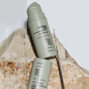 Zwei beige Flaschen mit NERDS. 0.1% Retinal-Serum stehen auf einer strukturierten Steinoberfläche, begleitet von einem Metall-Applikator.