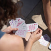 Zwei Personen spielen mit bunten Spielkarten im Freien. Die Karten zeigen kreative Symbole statt klassischer Motive wie Herzen oder Pik.