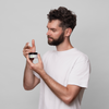 Ein Mann in einem weißen T-Shirt hält und öffnet ein Glas Scandinavian Biolabs Flexible Hybrid Clay und betrachtet es vor einem schlichten hellen Hintergrund.