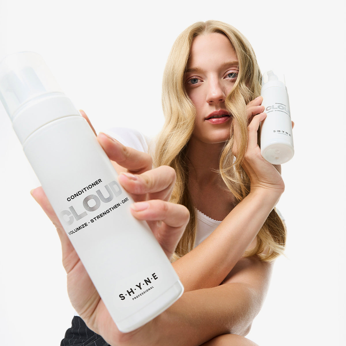 Eine Frau mit blondem Haar hält und präsentiert zwei Flaschen SHYNE Cloud Conditioner vor einem weißen Hintergrund.