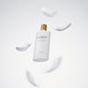 Madagascar Centella Air-Fit Sonnencreme Plus in einer weißen Flasche mit goldenem Verschluss, umgeben von schwebenden weißen Federn auf hellem Hintergrund.
