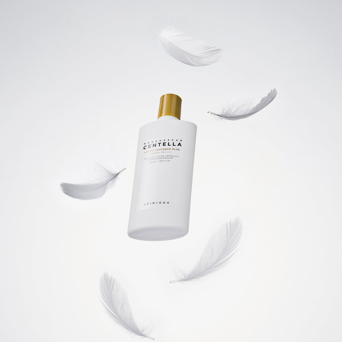 Madagascar Centella Air-Fit Sonnencreme Plus in einer weißen Flasche mit goldenem Verschluss, umgeben von schwebenden weißen Federn auf hellem Hintergrund.