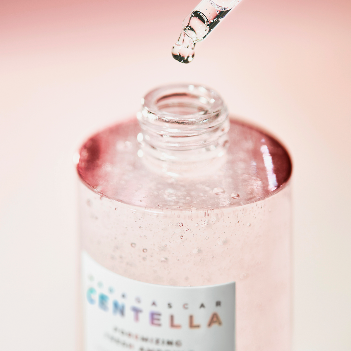 Nahaufnahme einer geöffneten Flasche mit rosa Ampulle. Eine Pipette mit einem Tropfen schwebt darüber. Das Etikett zeigt „CENTELLA“.