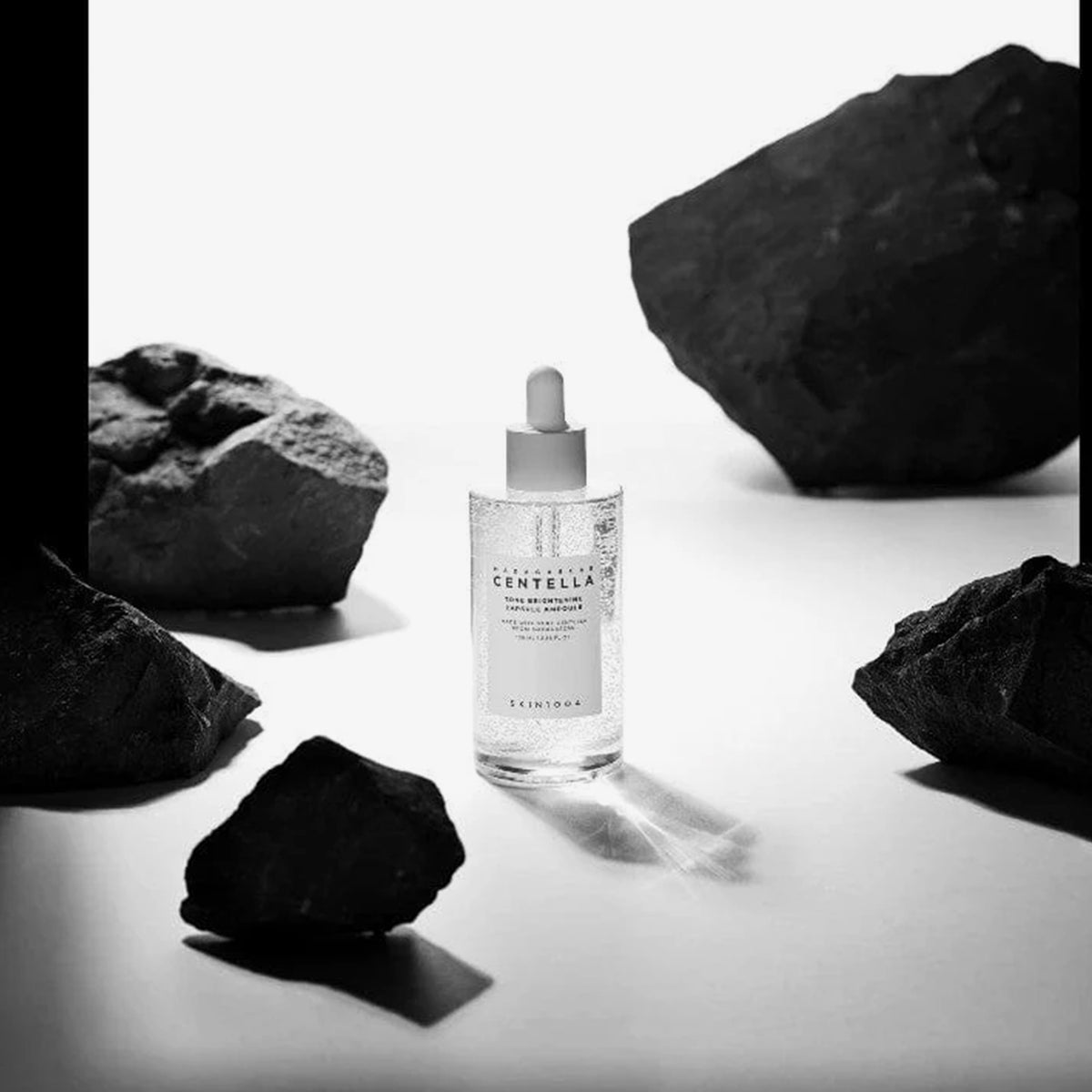 Die Madagascar Centella Tone Brightening Capsule Ampoule steht zwischen dunklen Felsen auf einer weißen Oberfläche und wird von dramatischer Beleuchtung erhellt.