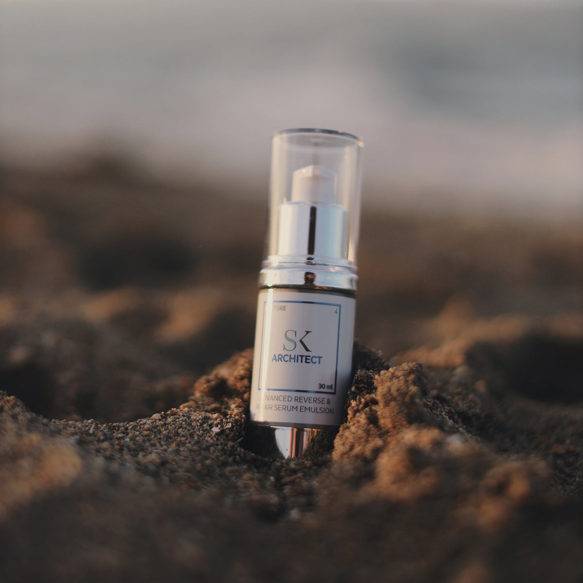 Eine Flasche Architect-Serum steht auf sandigem Boden vor einer verschwommenen Strandkulisse.