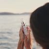 Eine Frau hält eine Cica-Cera-Hautpflegeflasche mit dem Meer und Sonnenuntergang im Hintergrund.