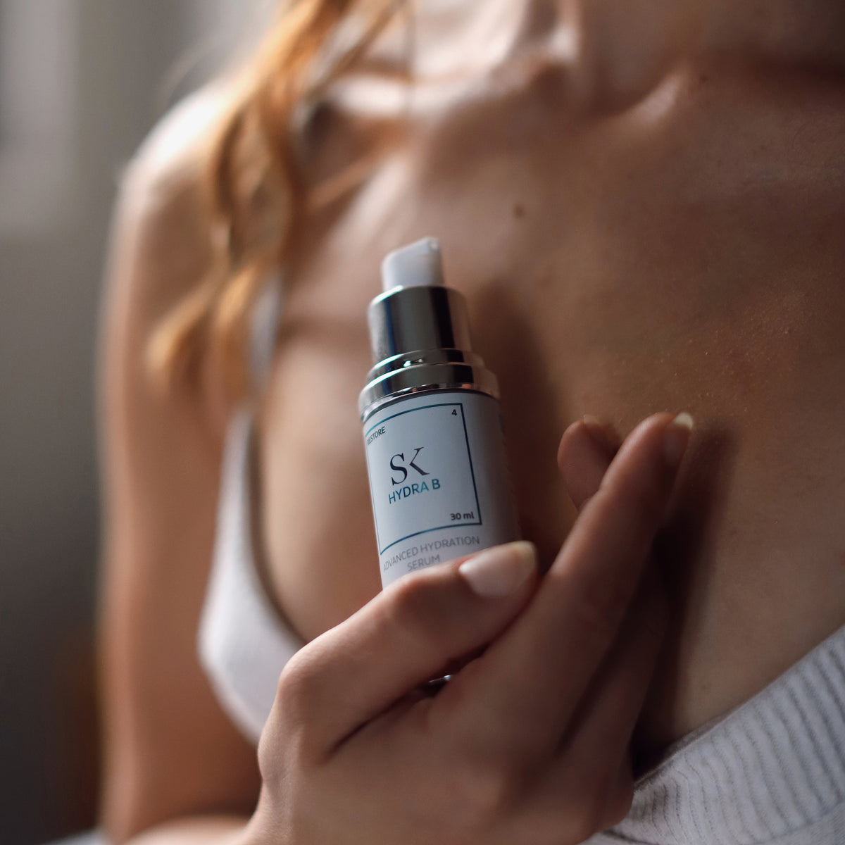 Frau in weißem Top hält eine Flasche Skintegra Hydra B Serum vor ihr Dekolleté in weichem, natürlichem Licht.