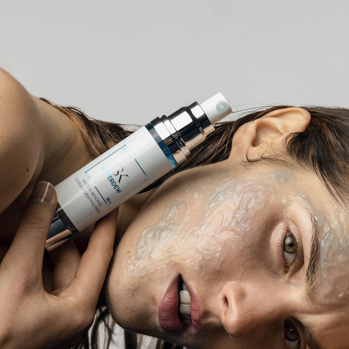 Eine FRau hält eine Hyperdew-Hautpflegeflasche an ihre Wange, während eine gelartige Maske auf ihr Gesicht aufgetragen ist.
