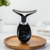 Der Fabulous Firmer Neck & Face Smoother in Schwarz und Silber liegt auf einem Holztablett, im Hintergrund sind ein Handtuch und eine Pflanze zu sehen.