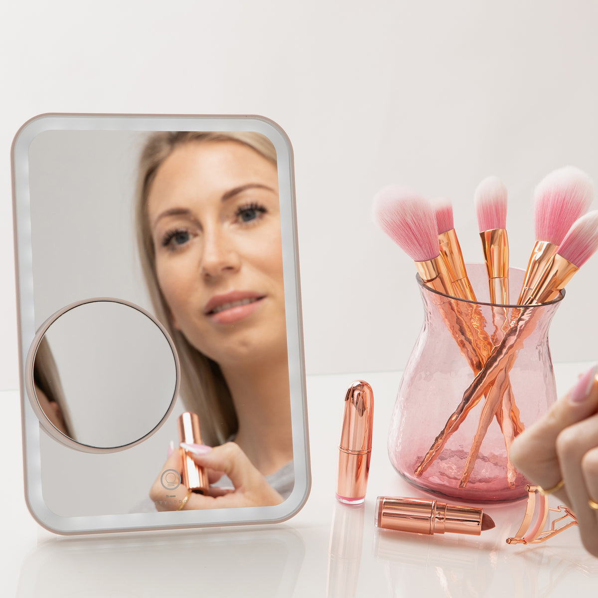 Eine Frau verwendet den Glow & Go Reisespiegel, um Lippenstift aufzutragen, während rosa Make-up-Pinsel in einem Glashalter in der Nähe ruhen.