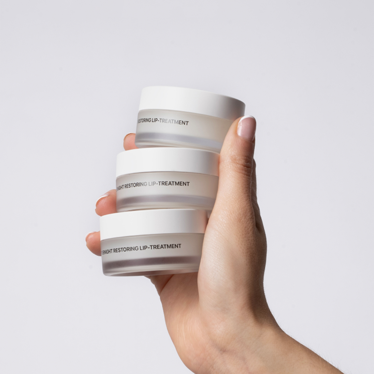 Eine Hand hält drei Overnight Restoring Lip-Treatments von Transparent Lab vor einem grau-weißen Hintergrund.