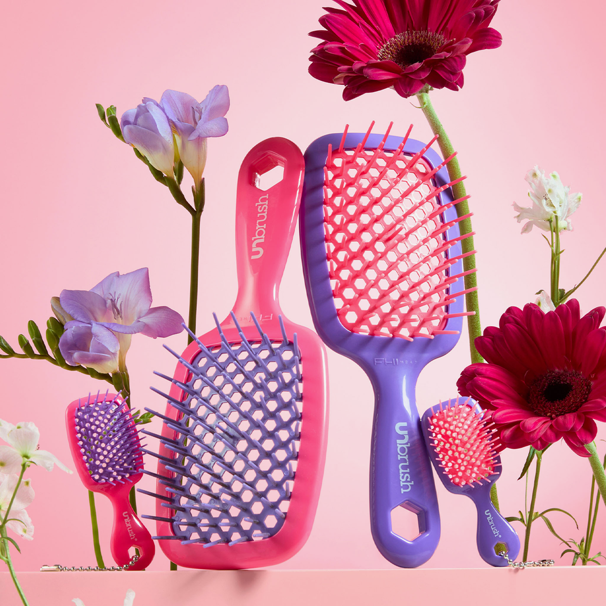 Zwei neonviolette und zwei neonrosa UNbrush- und Super-Mini-Set-Haarbürsten in Neonviolett, arrangiert zwischen rosa und violetten Blumen.