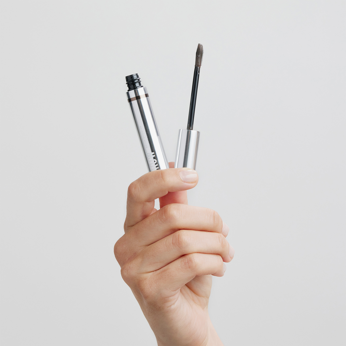 Eine Hand hält eine geöffnete Tube des XLASH Brow Booster-Augenbrauengels mit einem Applikator in Mascara-Optik.